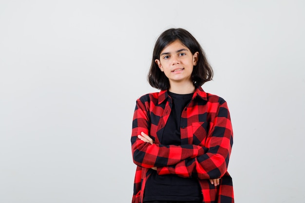 Ragazza teenager in piedi con le braccia incrociate in maglietta, camicia a scacchi e sembra soddisfatta. vista frontale.