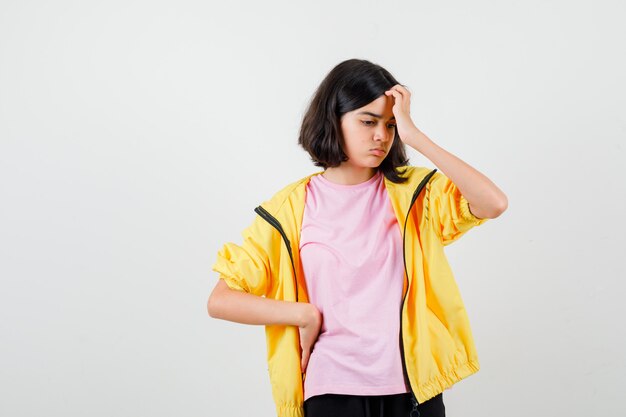 Ragazza teenager in t-shirt, giacca che si gratta la testa e sembra pensierosa, vista frontale.