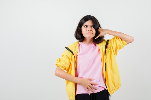Ragazza teenager in t-shirt, giacca che tiene la mano sullo stomaco, mostra il gesto del telefono e sembra esitante, vista frontale.