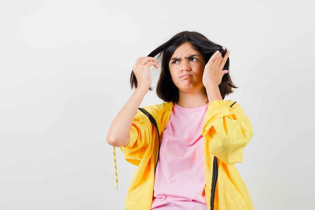 Ragazza teenager in tuta gialla, t-shirt che si tiene per mano nei capelli e sembra triste, vista frontale.