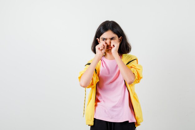 Ragazza teenager in tuta gialla, t-shirt stropicciandosi gli occhi con i pugni mentre piange e sembra cupa, vista frontale.