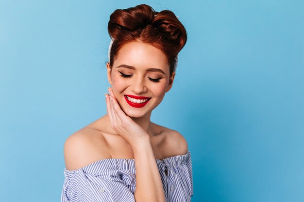 Ragazza timida dello zenzero che sorride con gli occhi chiusi. Studio shot di emotiva donna pinup in camicetta blu.