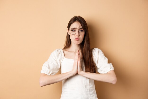 Ragazza triste con gli occhiali che implora aiuto dire per favore e sembra carina ha bisogno di un favore in piedi su uno sfondo beige.