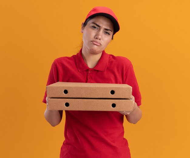 Ragazza triste di consegna giovane che indossa l'uniforme e cappuccio che tiene le scatole per pizza isolate sulla parete arancione