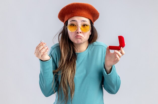 Ragazza triste il giorno di San Valentino che indossa un cappello con gli occhiali che tengono l'anello nuziale isolato su sfondo bianco