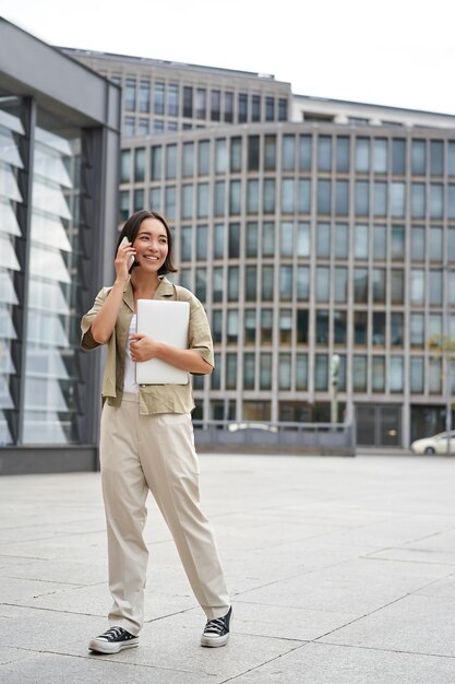 Ragazza urbana alla moda che cammina sulla via che parla sul telefono cellulare e che porta la fabbricazione della giovane donna del computer portatile