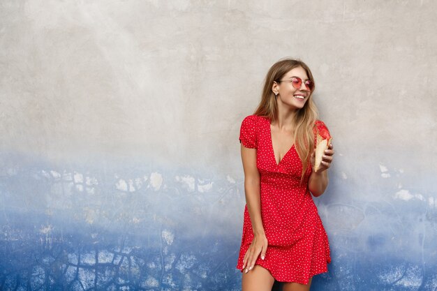 Ragazza urbana alla moda in occhiali da sole rossi e abito estivo, mangiare il gelato all'aperto, appoggiarsi a un muro di cemento e distogliere lo sguardo