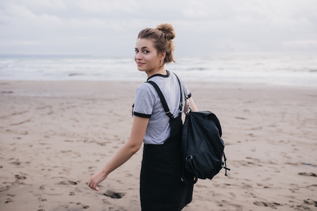 Ragazza vaga con zaino nero che osserva sopra la spalla mentre si cammina lungo la costa. Foto all'aperto del modello femminile bianco allegro che passa il tempo in spiaggia.