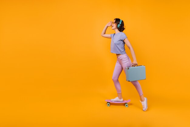 Ragazza vaga in pantaloni rosa in piedi su skateboard e ascolto di musica. Modello femminile riccio ispirato in cuffie in posa con valigia blu.