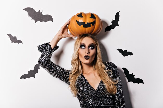 Ragazza vampiro dagli occhi azzurri in piedi sul muro bianco con i pipistrelli. Tiro al coperto di signora bionda interessata con zucca di Halloween.