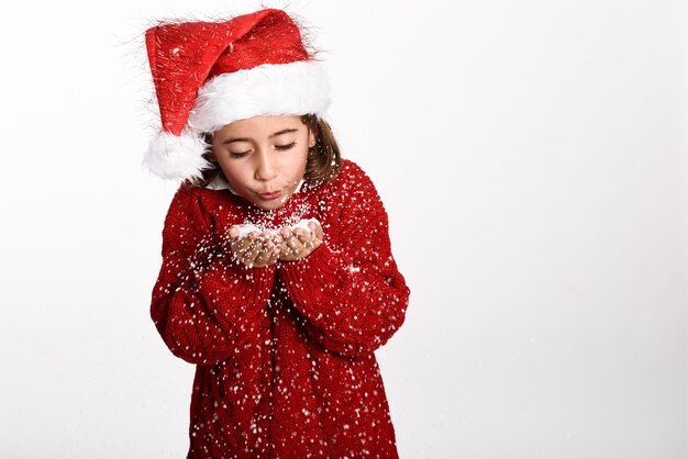 Ragazza vestita con abiti invernali e cappello di Babbo Natale soffia fiocchi di neve dalle sue mani su sfondo bianco