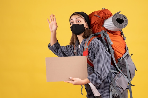 ragazza viaggiatrice con maschera nera e zaino con cartone che saluta qualcuno