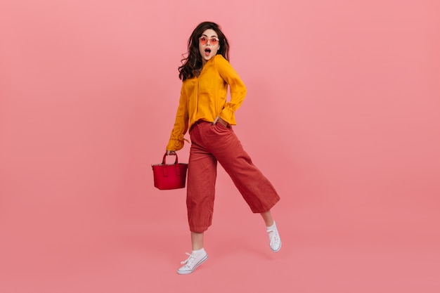 Ragazza vivace con gli occhiali alla moda guarda con stupore, camminando sul muro rosa. Bruna in culottes e camicetta arancione in posa con la borsa rossa.