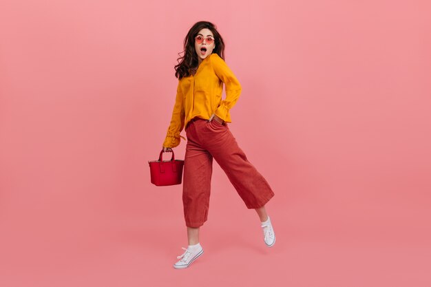 Ragazza vivace con gli occhiali alla moda guarda con stupore, camminando sul muro rosa. Bruna in culottes e camicetta arancione in posa con la borsa rossa.