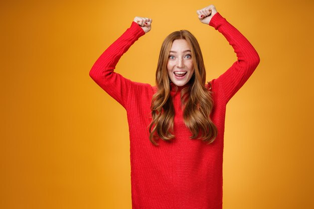 Ragazza zenzero ottimista felice e solidale che urla parole incoraggianti, alzando le mani con gioia e sorridendo ampiamente trionfante, celebrando il successo e la vittoria, posando soddisfatta ed eccitata contro il muro arancione