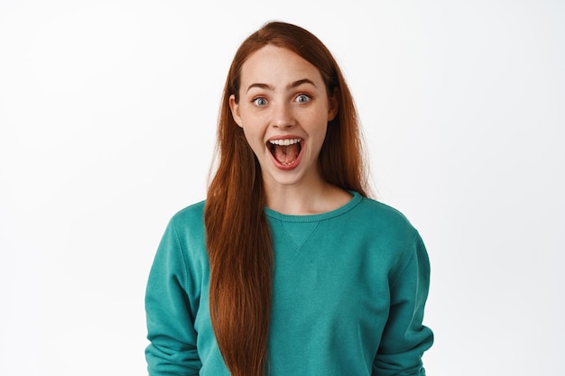 Ragazza zenzero sorpresa e felice, guarda con soggezione, guardando qualcosa di straordinario e fresco, sorridendo e fissando in soggezione, in piedi con una camicetta verde su sfondo bianco.