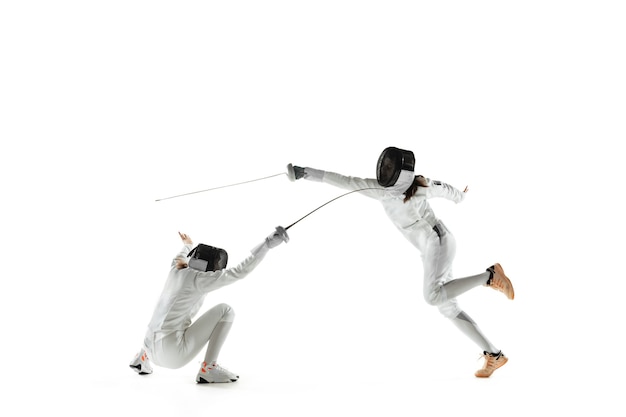 Ragazze adolescenti in costumi di scherma con le spade nelle mani isolate su sfondo bianco studio. Giovani modelli femminili che praticano e si allenano in movimento, azione. Copyspace. Sport, gioventù, stile di vita sano.