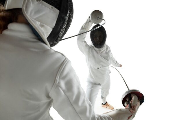 Ragazze adolescenti in costumi di scherma con le spade nelle mani isolate su sfondo bianco studio. Giovani modelli femminili che praticano e si allenano in movimento, azione. Copyspace. Sport, gioventù, stile di vita sano.