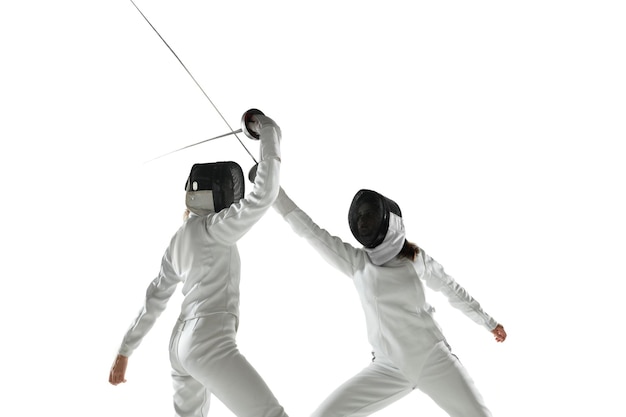 Ragazze adolescenti in costumi di scherma con le spade nelle mani isolate su sfondo bianco studio. Giovani modelli femminili che praticano e si allenano in movimento, azione. Copyspace.