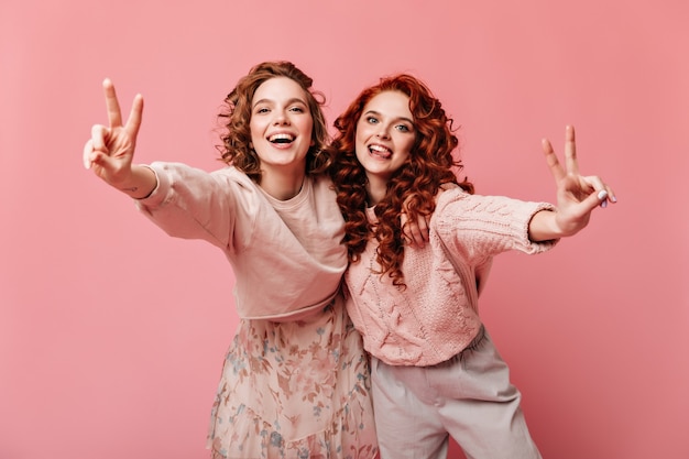 Ragazze adorabili che mostrano segni di pace e sorridenti. Studio shot di due amici isolati su sfondo rosa.