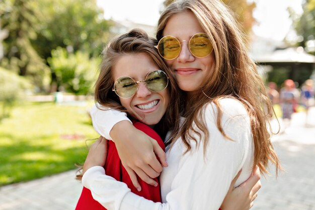 Ragazze amichevoli felici che si abbracciano con sorrisi veri e si divertono fuori. Ritratto di due signore eccitate che esprimono emozioni positive godendo di camminare.