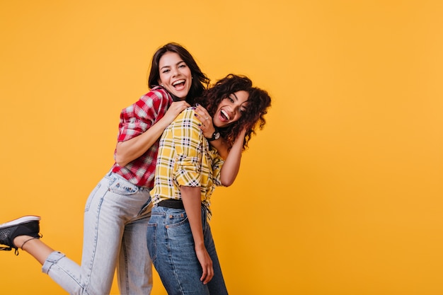 Ragazze attive ballano in jeans e camicette a quadretti. Le amiche si abbracciano e ridono sinceramente.