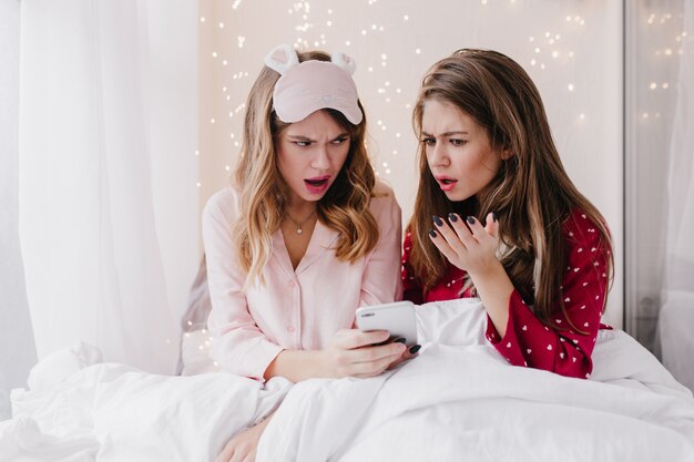 Ragazze caucasiche sconvolte che leggono il messaggio di telefono a letto. Foto al coperto di donne tristi dai capelli scuri che guardano lo schermo dello smartphone.