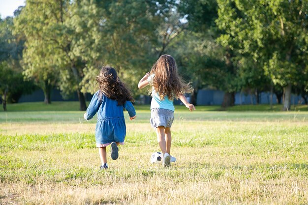 Ragazze dai capelli nere attive in esecuzione per pallone da calcio sull'erba nel parco cittadino. Lunghezza intera, vista posteriore. Infanzia e concetto di attività all'aperto