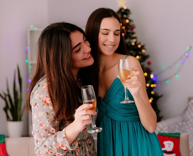 Ragazze graziose sorridenti che tengono bicchieri di champagne godendosi il periodo natalizio a casa at