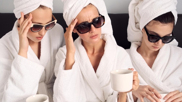 Ragazze sconvolte in accappatoi bianchi e occhiali da sole con tazze di caffè che hanno mal di testa dopo la festa nella camera d'albergo Espressione triste
