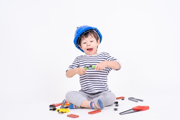 Ragazzino che indossa casco blu che si siede e che gioca con il giocattolo dell'attrezzatura per l'edilizia sul bianco