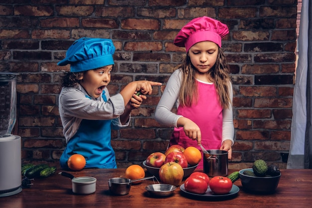 Ragazzino con capelli ricci castani vestito con un'uniforme da cuoco blu e una bella ragazza vestita con un'uniforme da cuoco rosa che cucinano insieme in una cucina contro un muro di mattoni. Piccola coppia carina di cuochi.