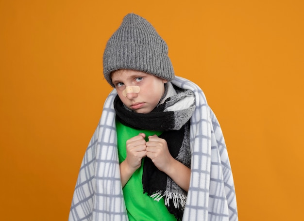 Ragazzino malato che indossa cappello e sciarpa caldi avvolti in una coperta con la toppa sul naso infelice e malato che osserva in piedi sopra la parete arancione