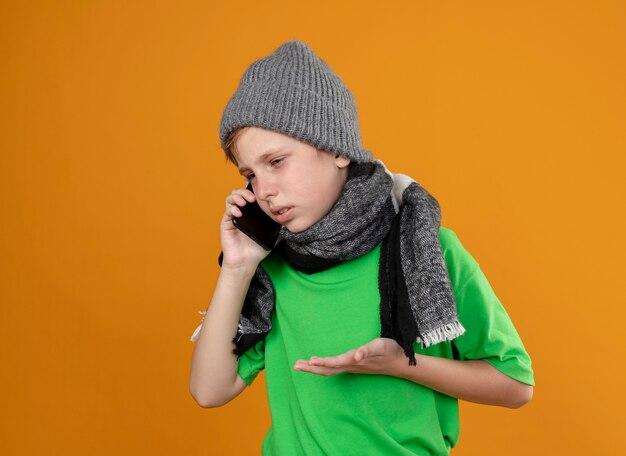 Ragazzino malato che indossa la maglietta verde in sciarpa calda e cappello sensazione di malessere parlando al telefono cellulare essendo scontento in piedi sopra la parete arancione