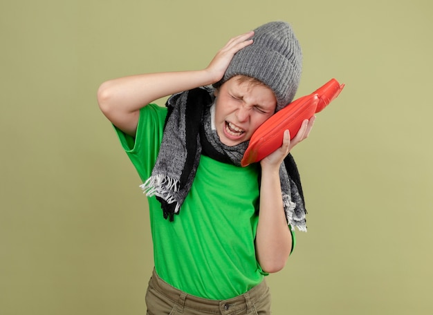 Ragazzino malato che indossa la maglietta verde in una sciarpa calda e cappello sensazione di malessere tenendo la bottiglia d'acqua per mantenere calda la sofferenza da forte mal di testa in piedi sopra la parete chiara