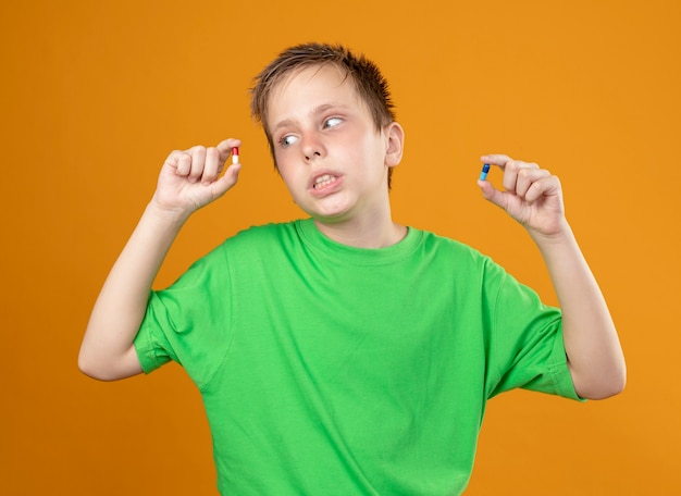 Ragazzino malato in maglietta verde che si sente male mostrando le pillole nelle mani che cercano confuso cercando di fare la scelta in piedi su sfondo arancione