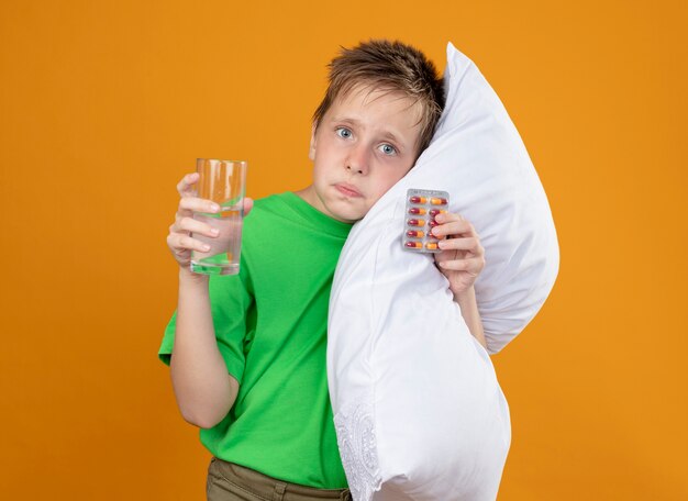 Ragazzino malato in maglietta verde che si sente male tenendo il cuscino e un bicchiere di acqua con le pillole che si appoggia la testa sul cuscino confuso in piedi sopra la parete arancione