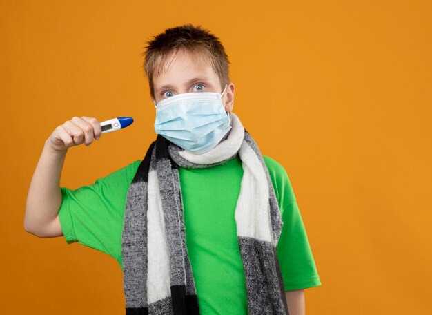 Ragazzino malato in maglietta verde e sciarpa calda intorno al suo collo maschera protettiva facciale che mostra il tremometro che sembra preoccupato in piedi sopra la parete arancione