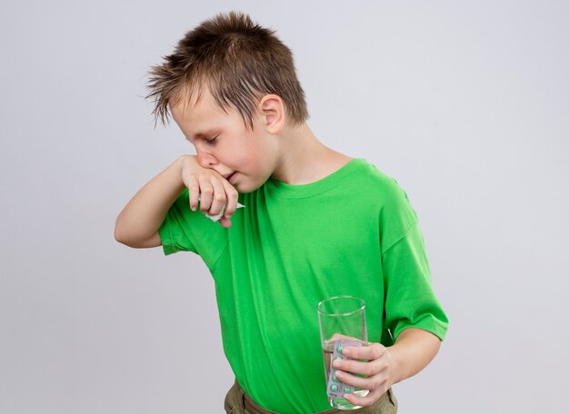 Ragazzino malato in maglietta verde sensazione di malessere tenendo un bicchiere di acqua e pillole pulendosi il naso con un tovagliolo di carta in piedi sopra il muro bianco