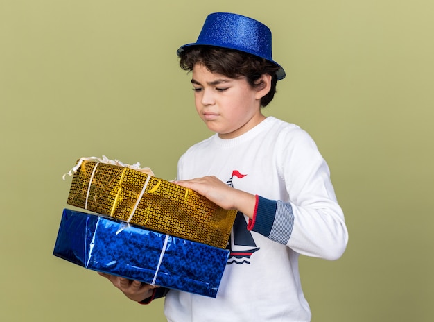 Ragazzino scontento che indossa un cappello da festa blu che tiene in mano e guarda scatole regalo isolate su una parete verde oliva