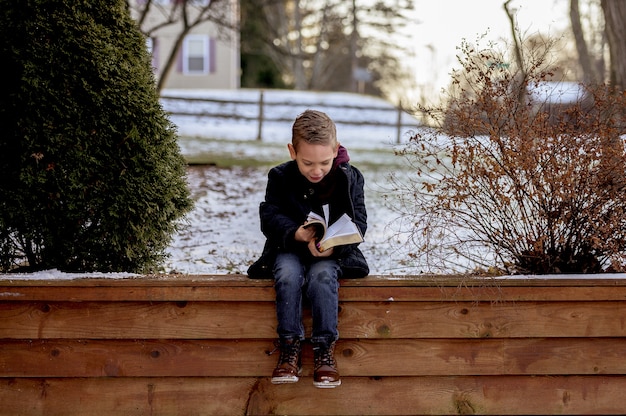 Ragazzino seduto su assi di legno e leggere la Bibbia in un giardino coperto di neve