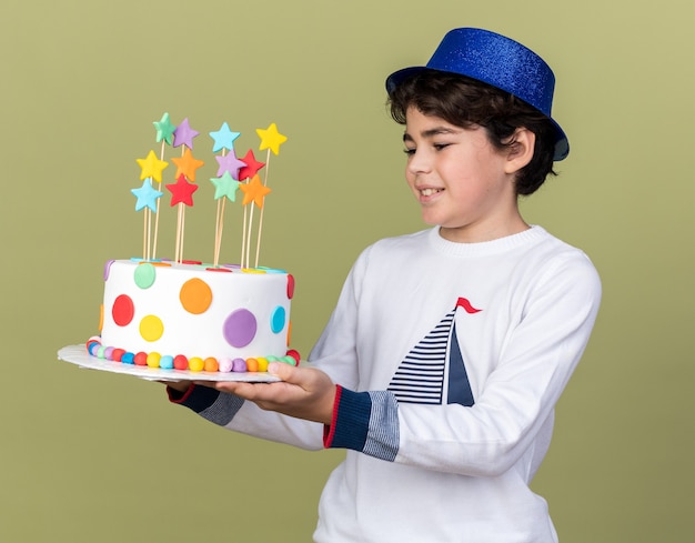 Ragazzino sorridente che indossa un cappello da festa blu che tiene in mano e guarda la torta