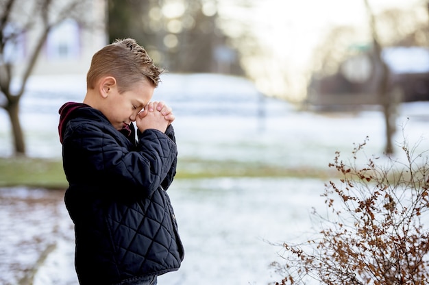 Ragazzino sveglio che prega con gli occhi chiusi nel mezzo del parco invernale