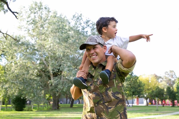 Ragazzino uscito seduto sul collo di papà e indicando lontano. Padre caucasico che tiene le gambe del figlio, sorridente, con indosso l'uniforme dell'esercito e passeggiate nel parco. Ricongiungimento familiare, paternità e concetto di ritorno a casa