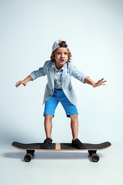 Ragazzo abbastanza giovane su skateboard in abiti casual sul muro bianco. Cavalca e sembra felice. Bambino in età prescolare maschio caucasico con emozioni facciali luminose. Infanzia, espressione, divertimento.