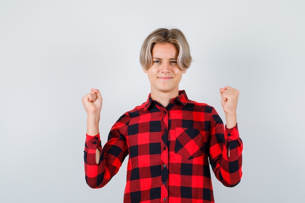 Ragazzo abbastanza teenager che mostra il gesto del vincitore in camicia a quadri e sembra fortunato. vista frontale.