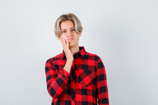 Ragazzo abbastanza teenager che si appoggia la guancia a portata di mano in camicia a quadri e sembra dispiaciuto, vista frontale.