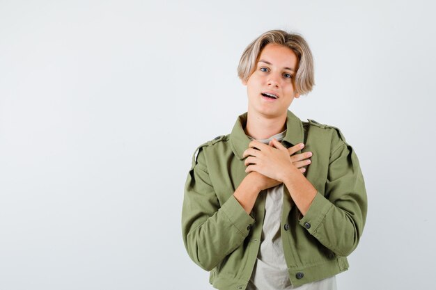 Ragazzo abbastanza teenager che tiene le mani sul petto in giacca verde e sembra grato. vista frontale.