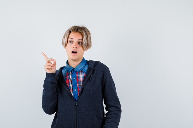 Ragazzo adolescente carino che punta all'angolo in alto a sinistra in camicia, felpa con cappuccio e sembra meravigliato. vista frontale.