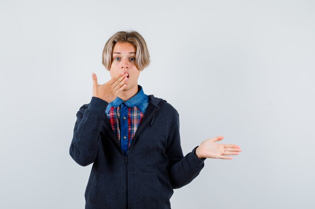 Ragazzo adolescente carino che tiene la mano sulla bocca, allarga il palmo in camicia, felpa con cappuccio e sembra sorpreso, vista frontale.